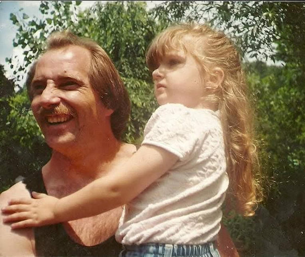 A vintage-style photograph showing a smiling man outdoors carrying a young girl on his shoulders. The man has light brown hair and a mustache, and the girl has long blonde hair and is wearing a white textured short-sleeve top.
